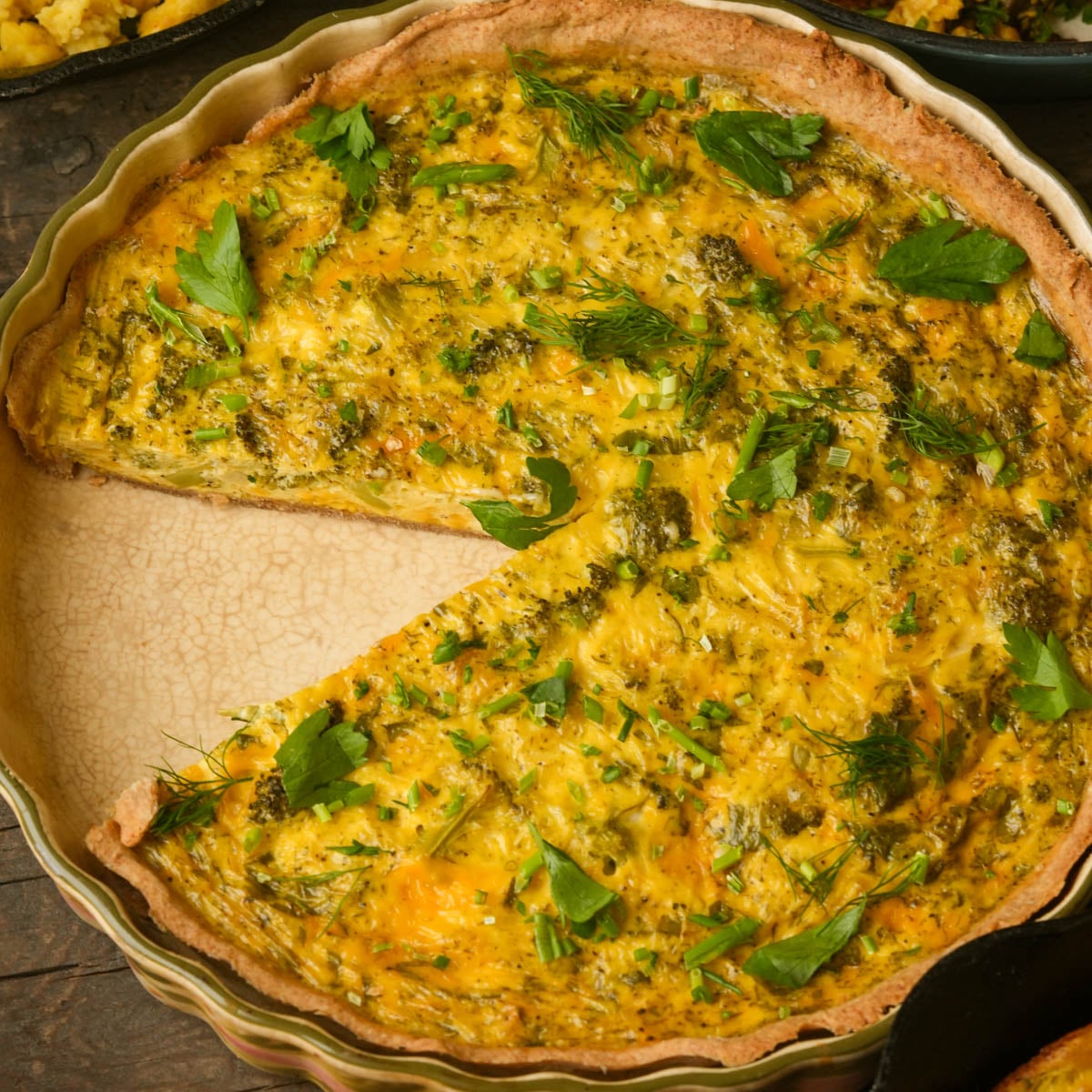 A baked vegan quiche with a slice missing, topped with fresh herbs and displayed in a round pie dish.