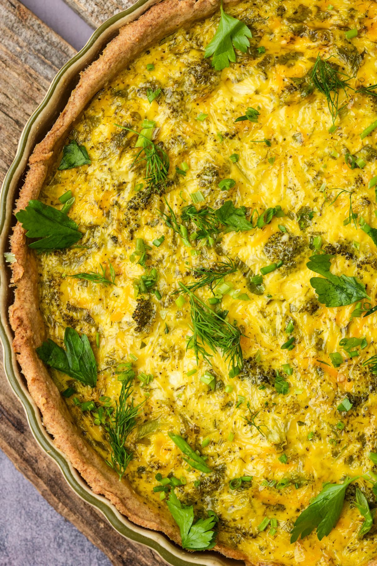 Close-up of a baked vegan quiche topped with fresh herbs, showing a golden crust and a textured egg filling with green vegetable pieces.