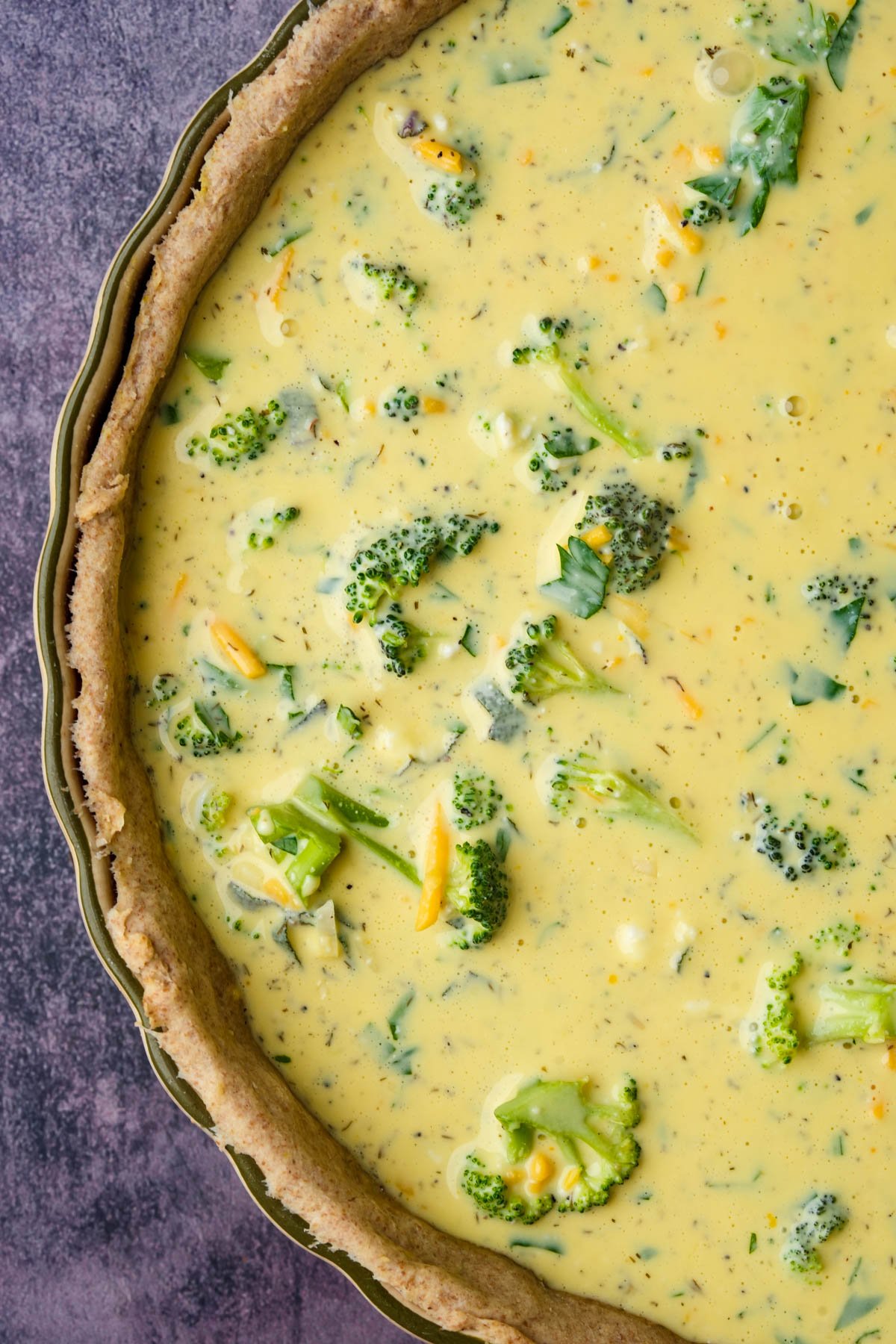 A close-up of an unbaked vegan quiche in a pie crust, filled with a yellow mung egg mixture and broccoli florets.