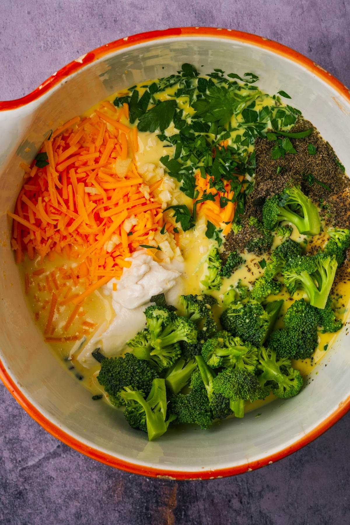 A mixing bowl containing shredded cheese, chopped parsley, seasonings, broccoli florets, a white creamy ingredient, and an mung egg mixture for making a vegan quiche.