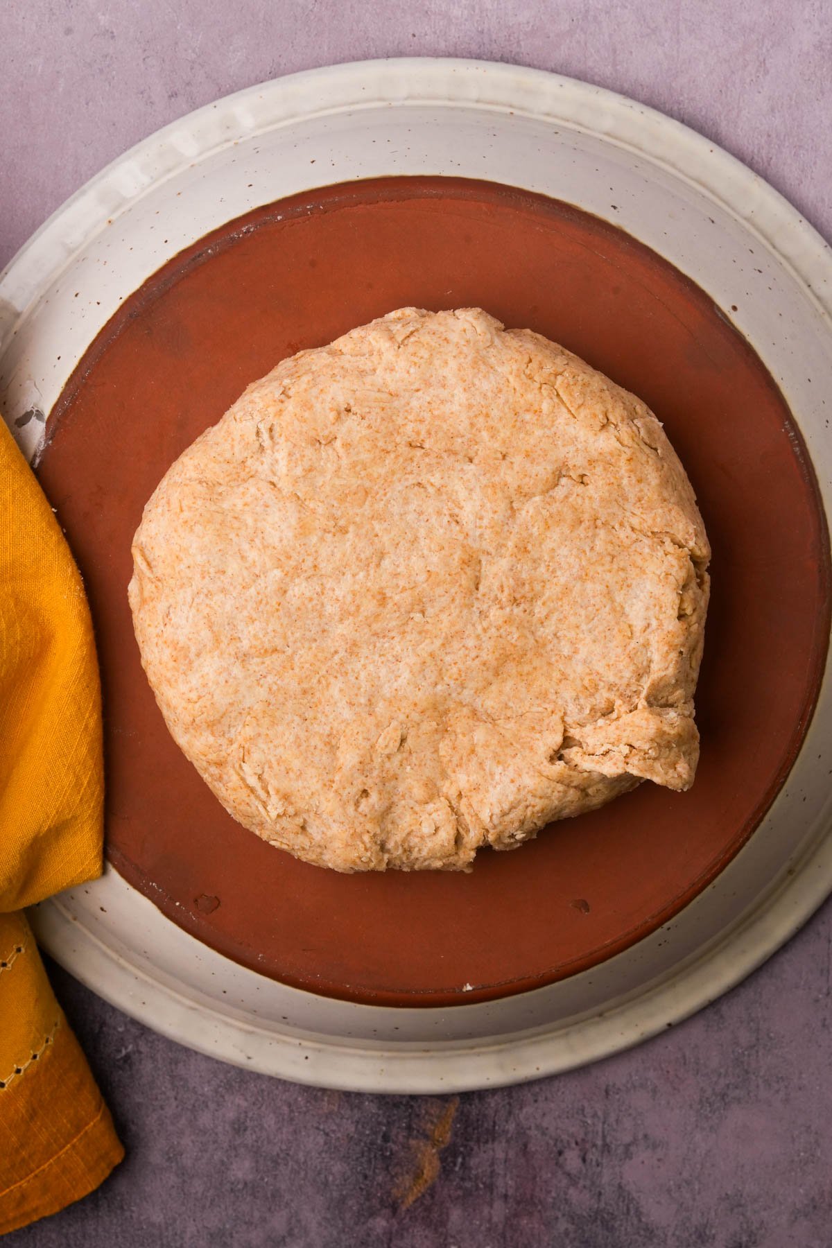 A round ball of dough sits on a brown baking stone placed on a white plate, with a yellow cloth partially visible to the side.