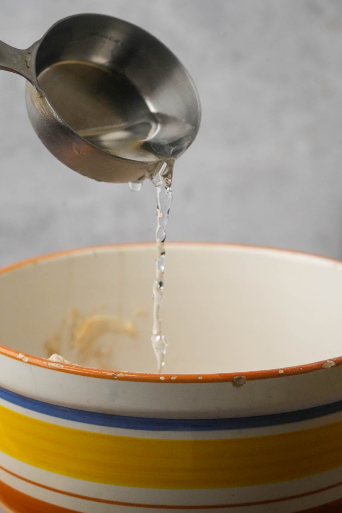 A metal measuring spoon pours cold water into a ceramic bowl with orange, blue, and yellow stripes.