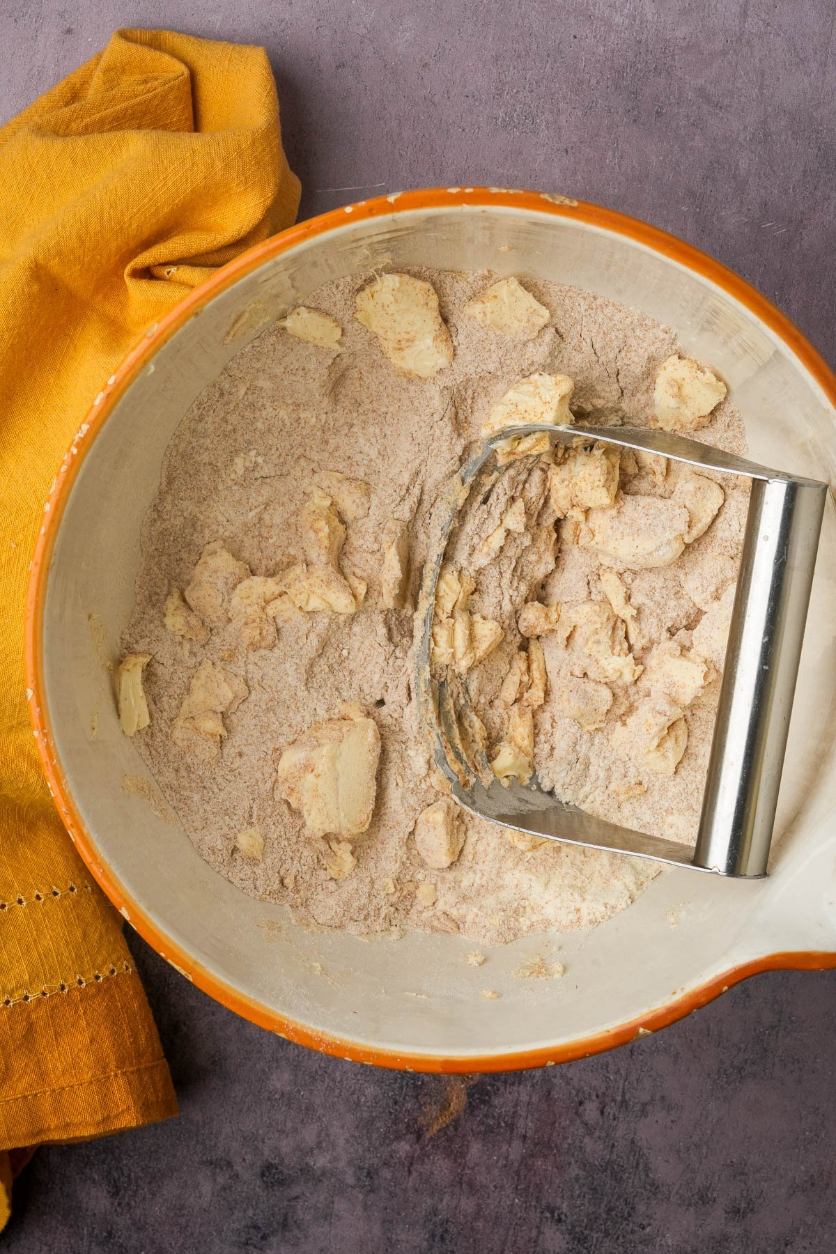 A mixing bowl with flour and butter being cut together using a pastry blender, next to a folded orange cloth on a gray surface.