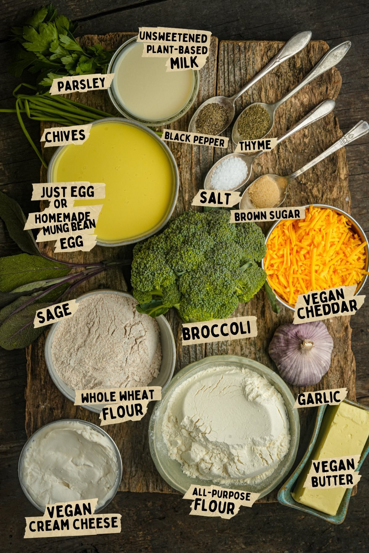Various labeled vegan quiche ingredients, including plant-based milk, flour, vegan cheddar, broccoli, chives, parsley, garlic, brown sugar, vegan butter, spices, and vegan cream cheese, arranged on a wooden surface.