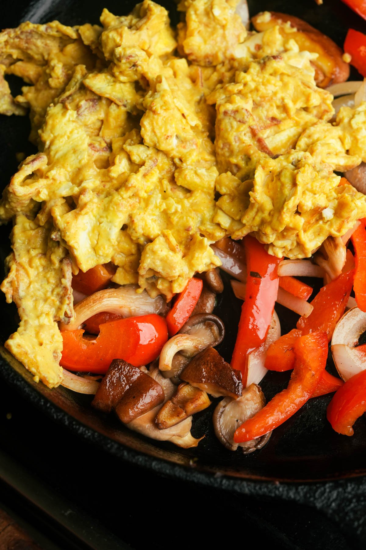 Vegan scrambled eggs, sliced red bell peppers, onions, and mushrooms cooking together in a black skillet.
