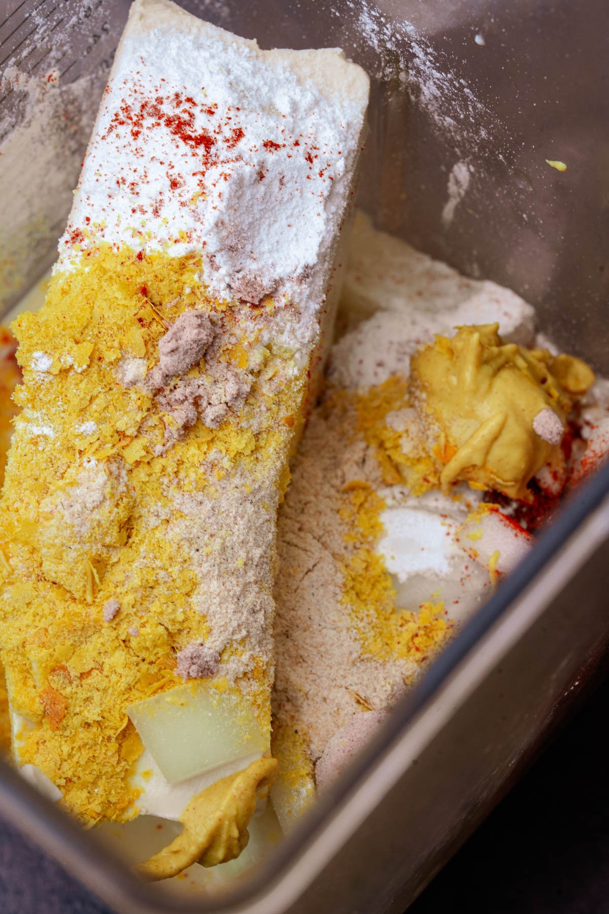 A close-up of various ingredients, including tofu, mustard, nutritional yeast, and powdered spices, in a blender.