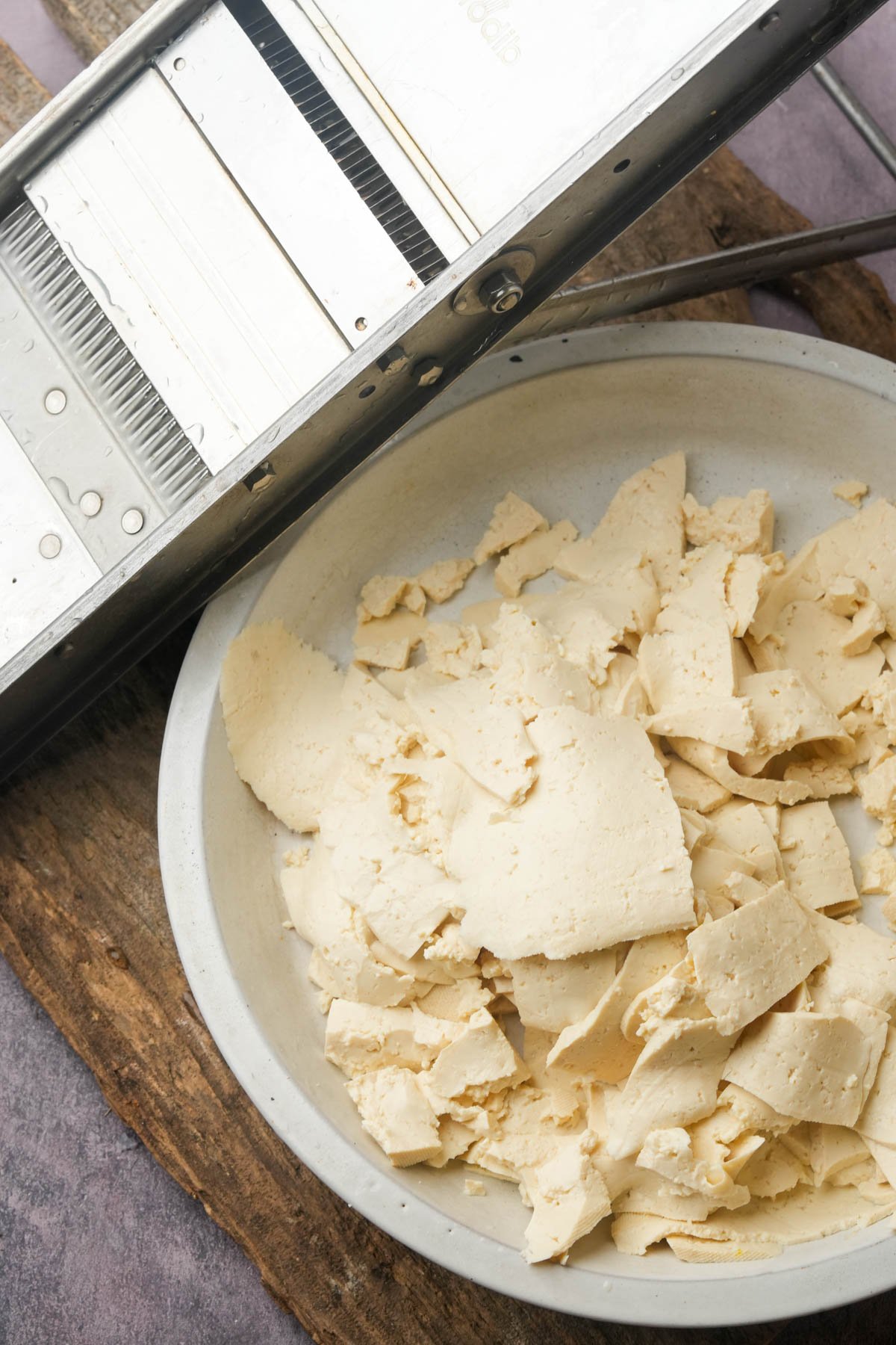 A white bowl filled with tofu slices sits on a wooden board next to a metal mandoline slicer.