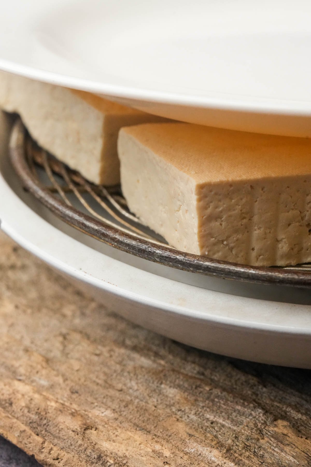 A plate presses water out of blocks of tofu placed on a wire rack over a dish.