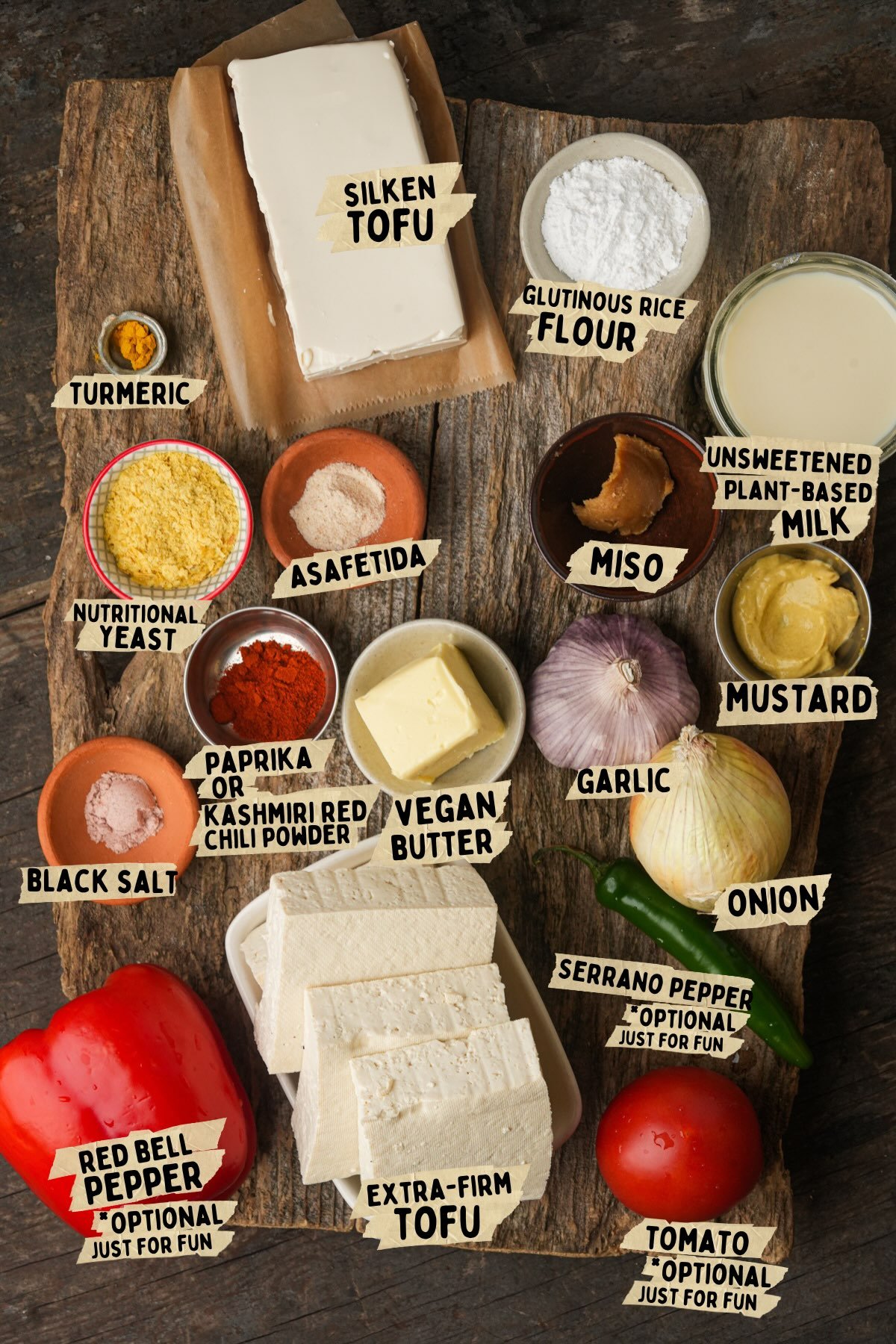 A wooden board displays ingredients for a vegan scrambled eggs recipe including silken and extra-firm tofu, rice flour, spices, miso, nutritional yeast, vegan butter, garlic, onion, pepper, and tomato.