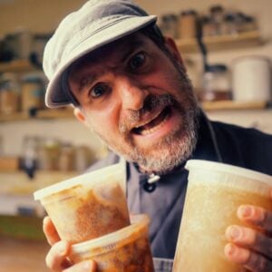 A man wearing a cap holds three plastic containers of food and looks intensely at the camera in a kitchen setting.