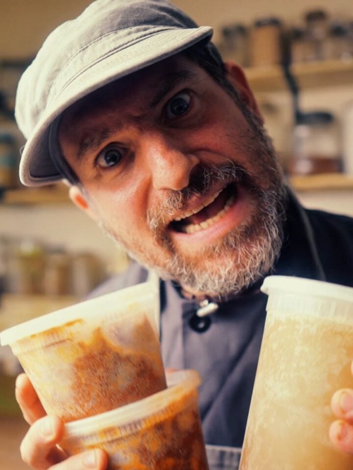 A man wearing a cap holds three plastic containers of food and looks intensely at the camera in a kitchen setting.