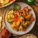 A plate of calabacitas with vegan cheese, corn, zucchini, tomatoes, and sliced peppers, served with salsa and tortillas on a wooden table.