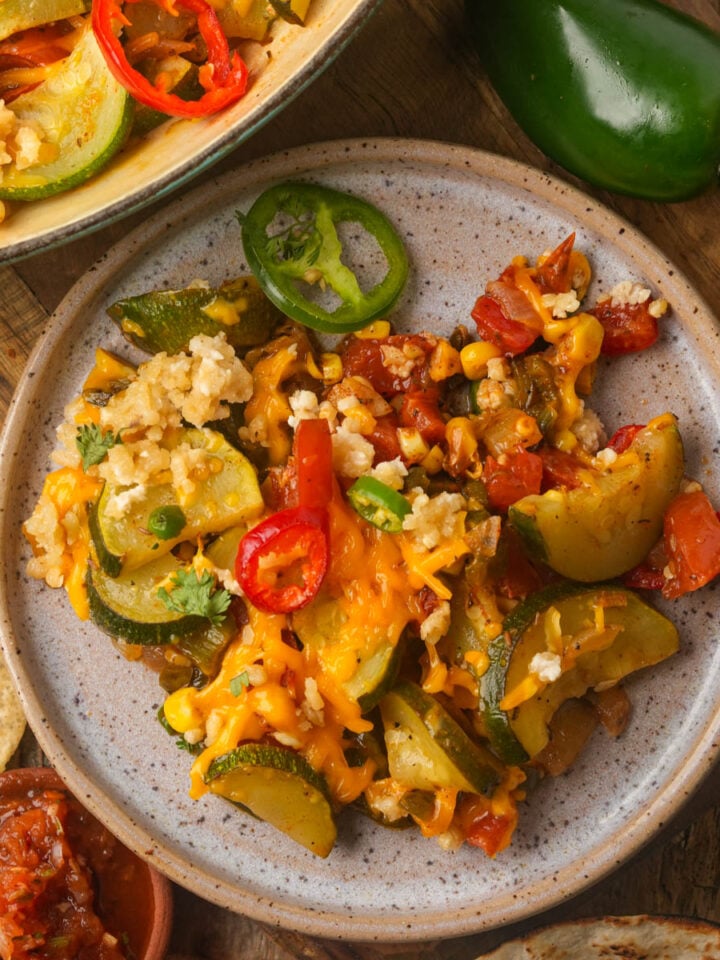 A plate of calabacitas topped with melted vegan cheese and sliced jalapeños, served with salsa and tortilla chips on a wooden table.