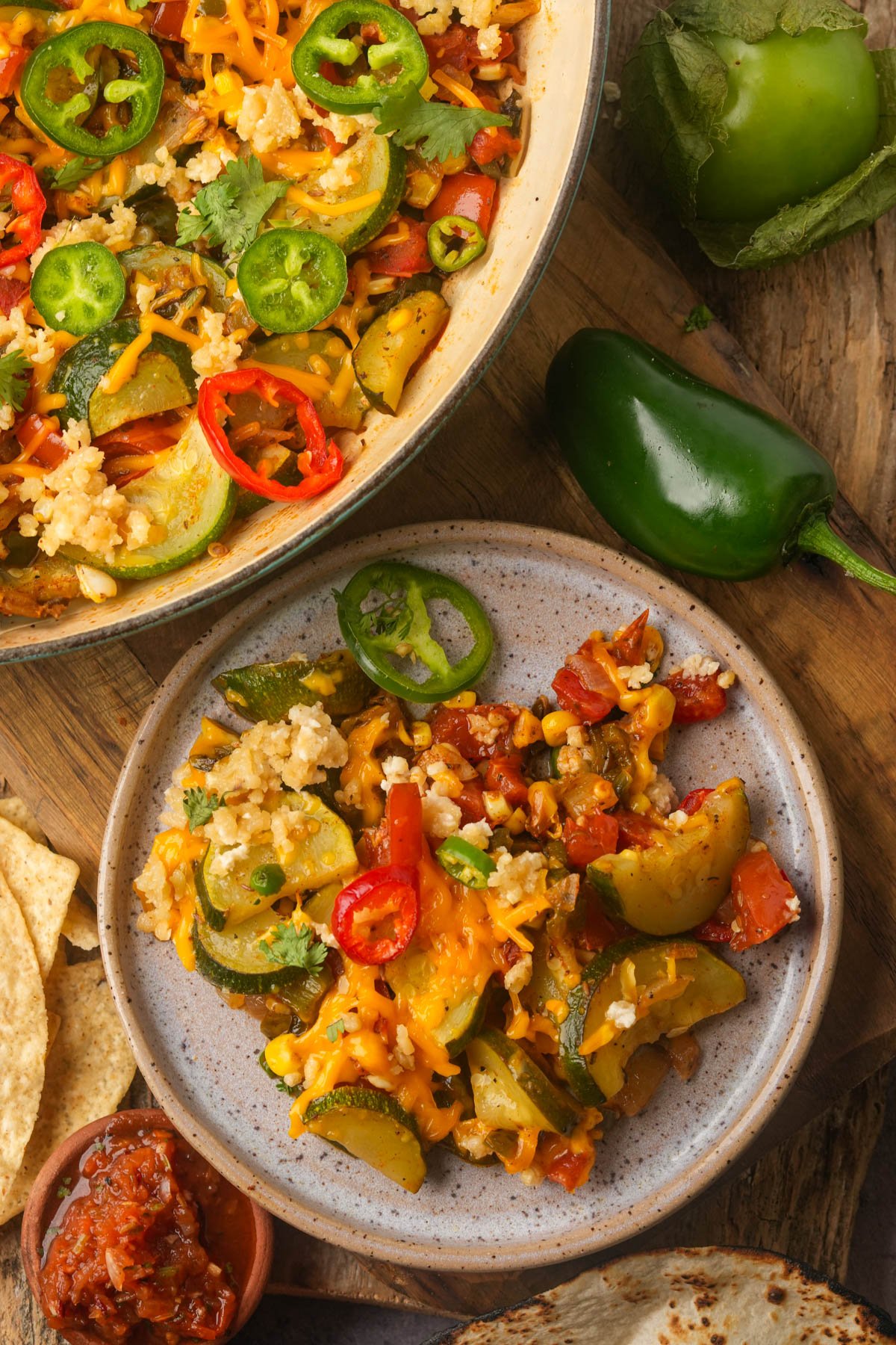 A plate and skillet with calabacitas topped with sliced peppers and vegan cheese, next to a green pepper, salsa, chips, and a tortilla on a wooden surface.