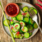 A plate of Filipino cucumber salad with red chili, shallots, and green onions, served with a small bowl of red chili dipping sauce and a fork on a rustic wooden surface.