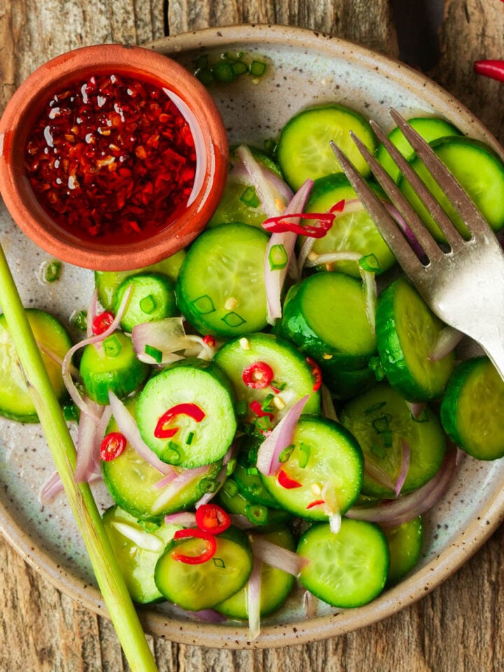 A plate of Filipino cucumber salad with red chili, shallots, and green onions, served with a small bowl of red chili dipping sauce and a fork on a rustic wooden surface.