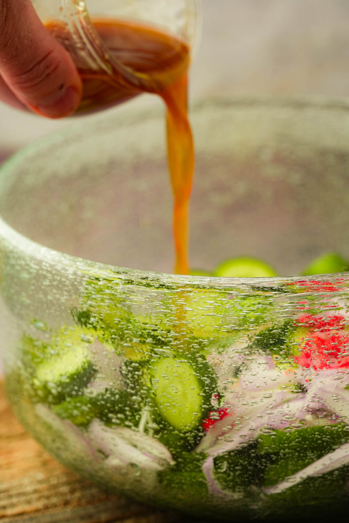 A hand pours brown dressing from a small glass container over a salad with cucumbers, onions, and chilies in a clear bowl.