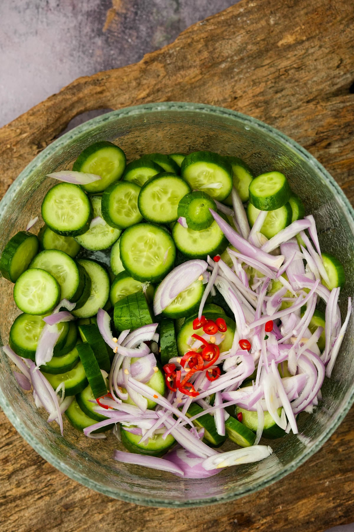 A glass bowl containing sliced cucumbers, thinly sliced red onions, and chopped red chili peppers on a wooden surface.