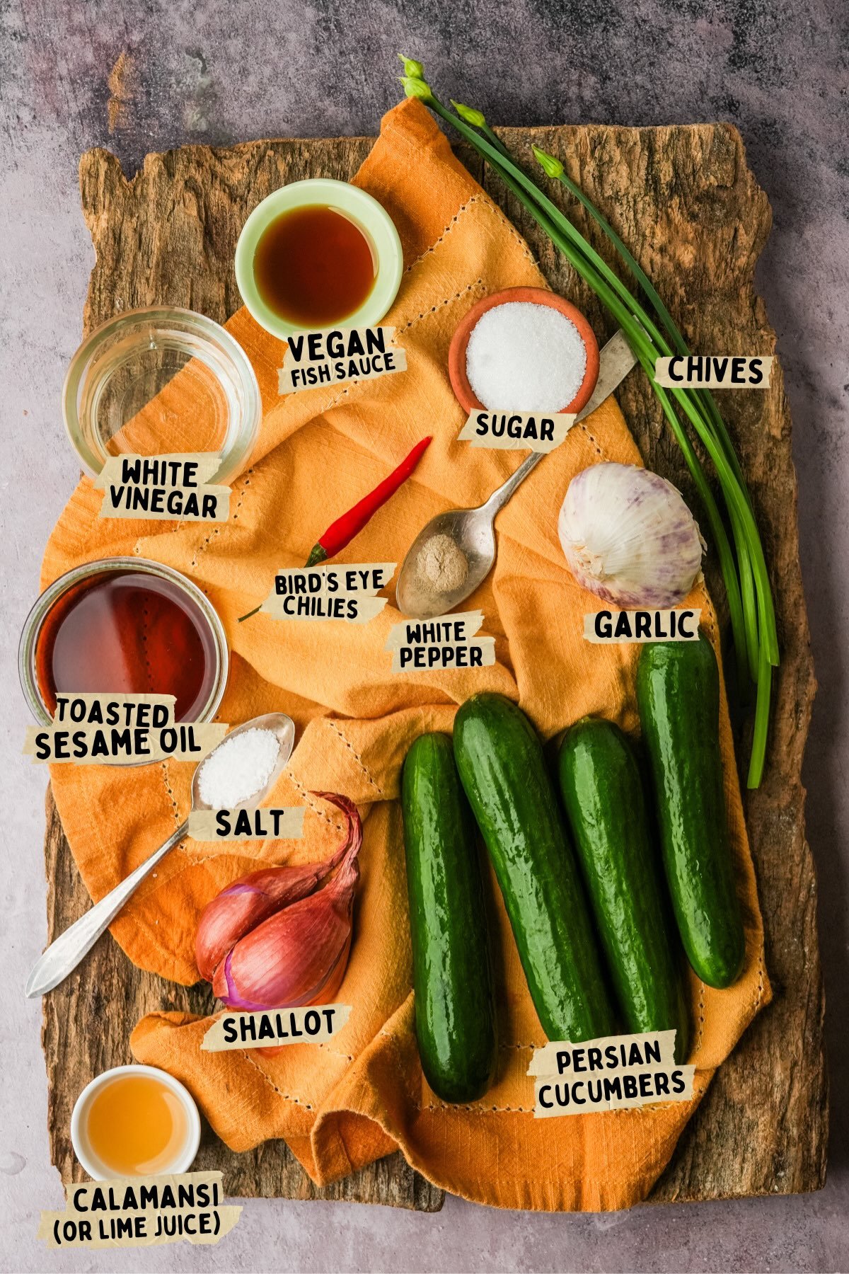 Ingredients for a Filipino cucumber salad (Ensaladang Pipino), including Persian cucumbers, chives, garlic, shallot, bird's eye chilies, sauces, spices, salt, sugar, vinegar, and lime juice, arranged on a wooden board.