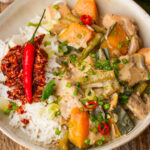 A bowl of rice topped with chili flakes and a red chili, served with ginataang gulay containing kalabasa, green beans, and sliced green onions.