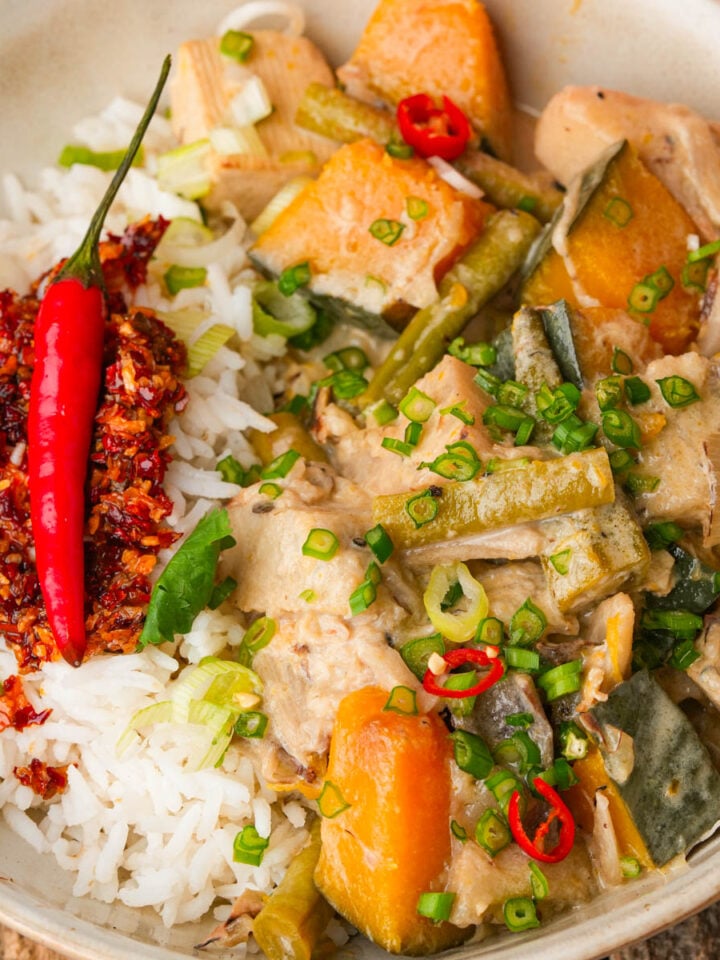A bowl of rice topped with chili flakes and a red chili, served with ginataang gulay containing kalabasa, green beans, and sliced green onions.