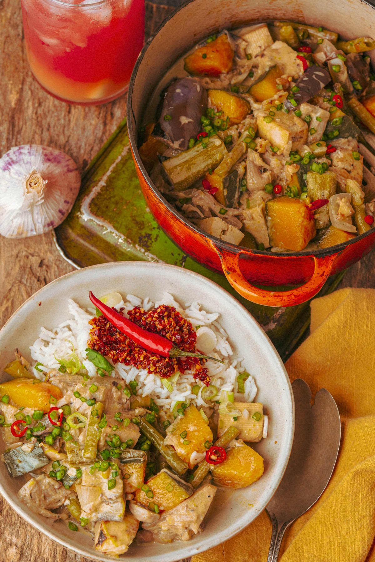 A bowl of rice topped with a ginataang gulay garnished with chopped green onions and red chilies, sits next to a pot of the same stew, a garlic bulb, and a glass of pink drink.