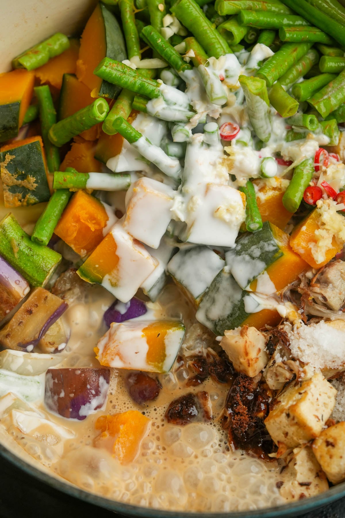 Chopped vegetables, including green beans, kalabasa, and eggplant, with coconut milk and seasonings in a pot, cooking.