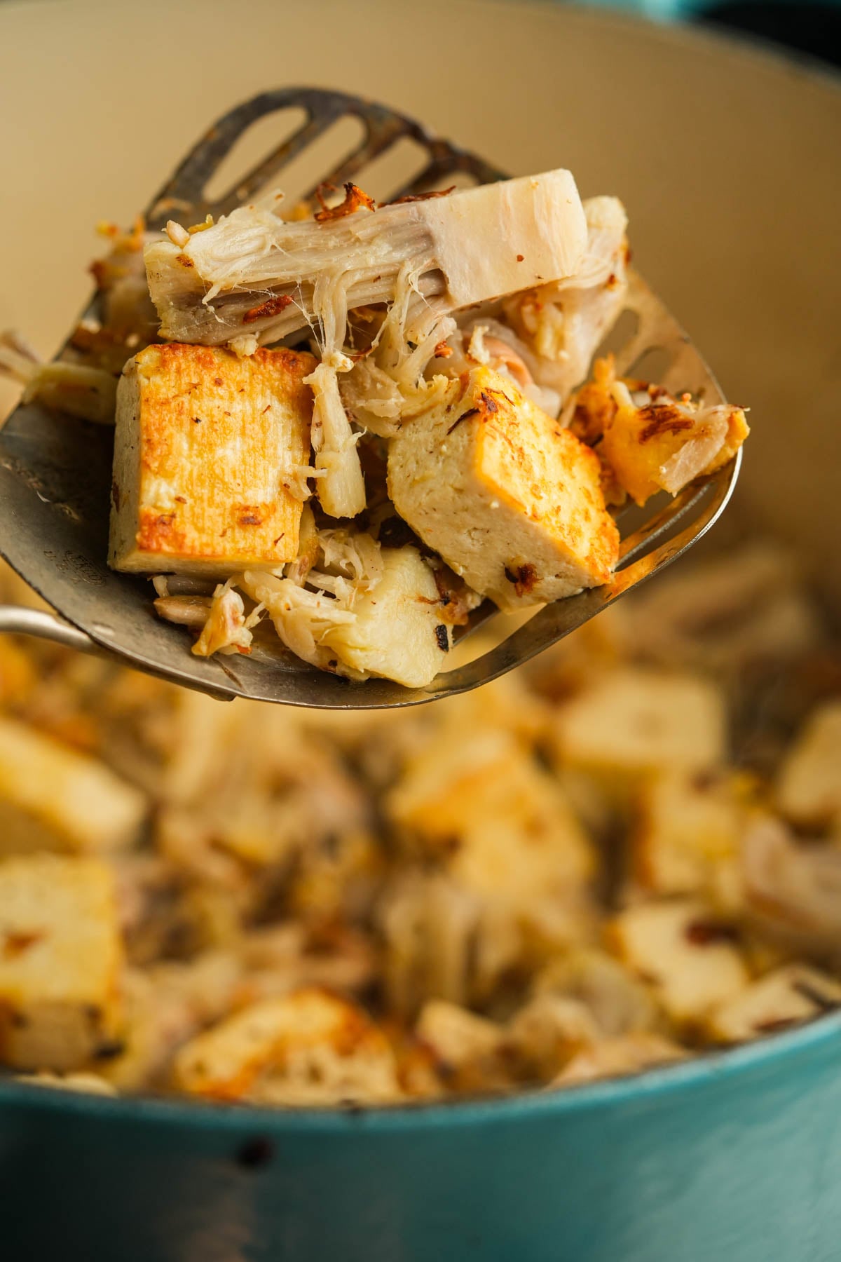 A slotted spatula holds a mix of browned tofu cubes and shredded jackfruit over a pot containing more of the same mixture.