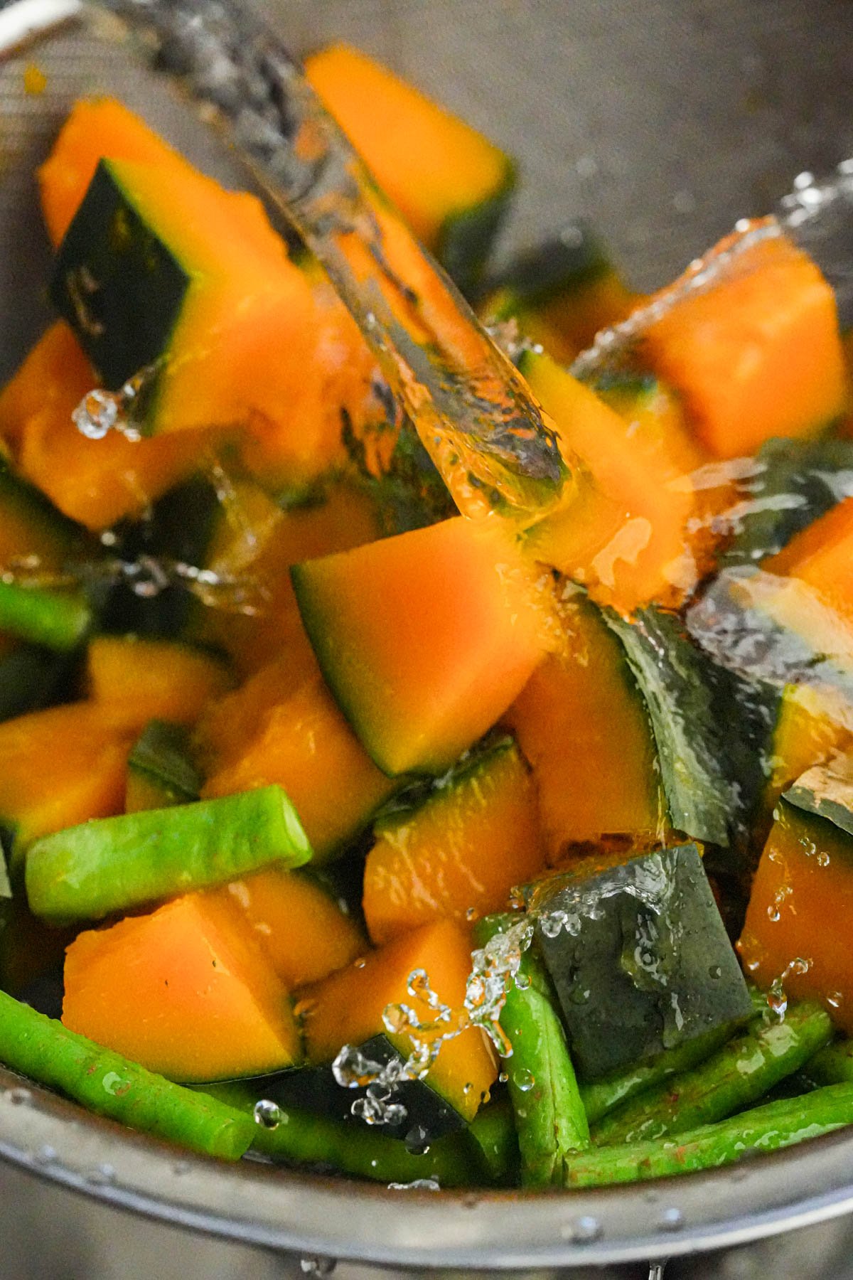 Chunks of cooked kalabasa and green beans are being rinsed with water in a metal colander.