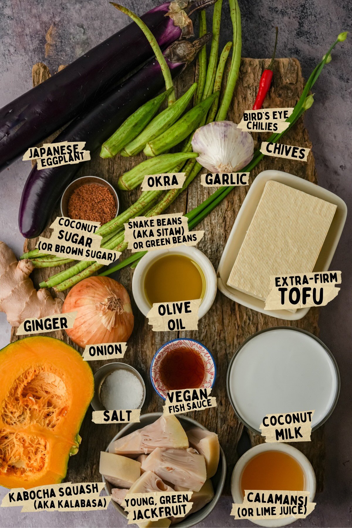 Ingredients for a ginataang gulau recipe displayed on a wooden surface, including Japanese eggplant, kabocha squash, tofu, spices, vegetables, oils, and condiments with labeled names.