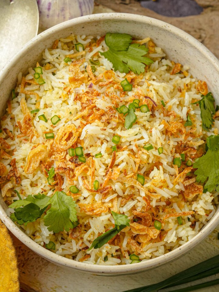 A bowl of cooked sinangag topped with fried shallots, chopped green onions, and fresh cilantro, placed on a rustic surface next to a yellow napkin and a metal spoon.