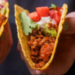 A hand holding a hard taco shell filled with seasoned ground tofu, chopped tomatoes, lettuce, avocado, onions, and vegan sour cream.