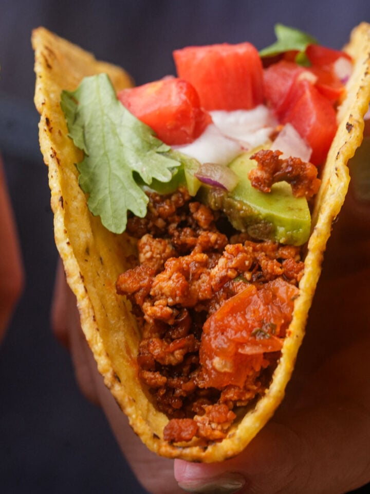 A hand holding a hard taco shell filled with seasoned ground tofu, chopped tomatoes, lettuce, avocado, onions, and vegan sour cream.