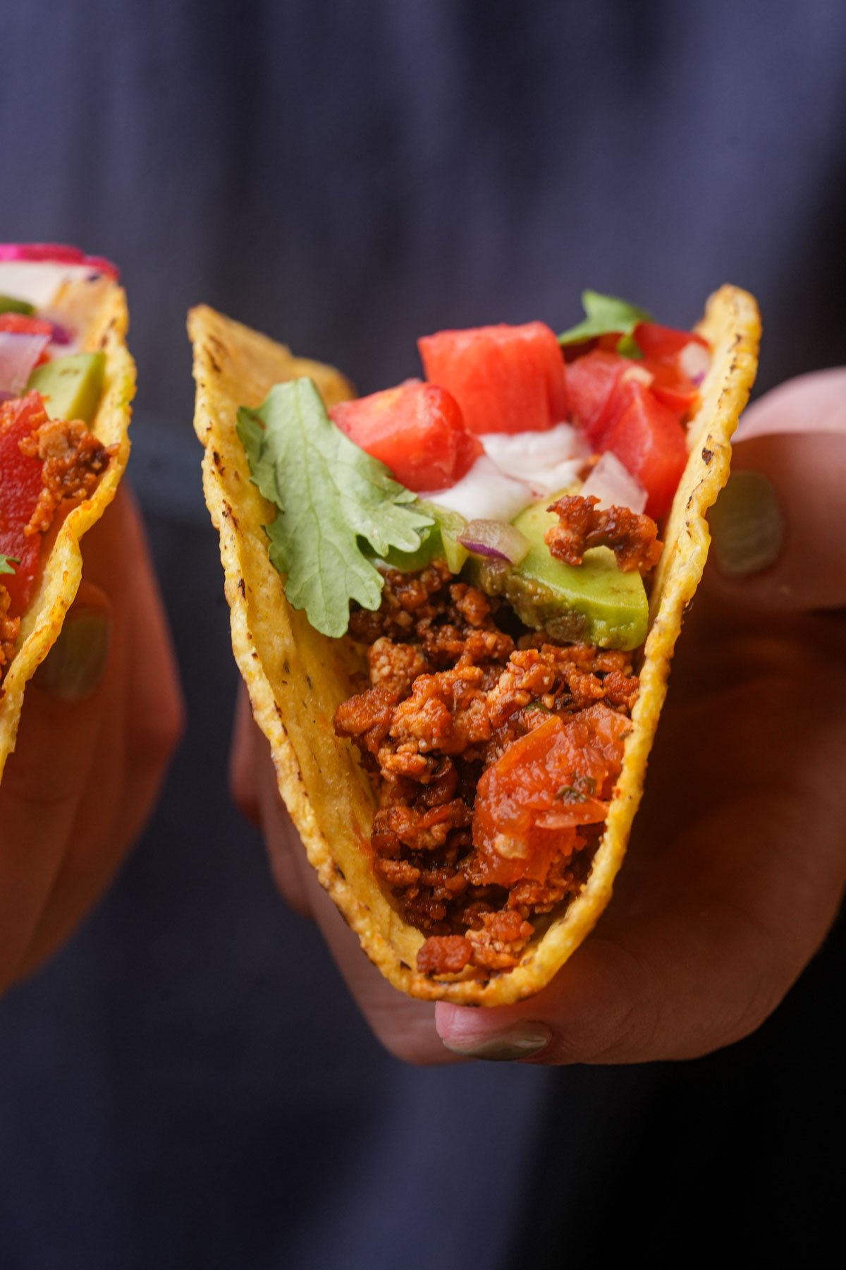 A hand holds a tofu taco filled with seasoned ground tofu, diced tomatoes, lettuce, guacamole, onions, and a dollop of vegan sour cream.