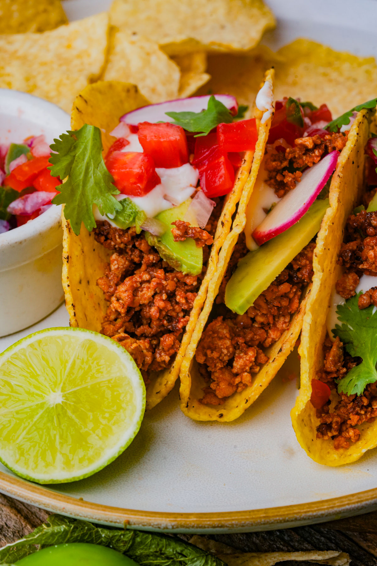 Three hard shell tacos filled with seasoned tofu taco meat, avocado slices, diced tomatoes, radishes, cilantro, and a creamy sauce, served with tortilla chips, salsa, and a halved lime.