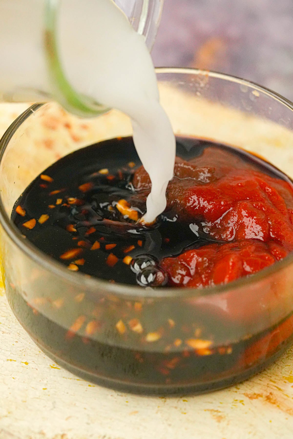A glass bowl contains tokwat baboy stir-fry sauce with a white cornstarch slurry being poured in from a glass.