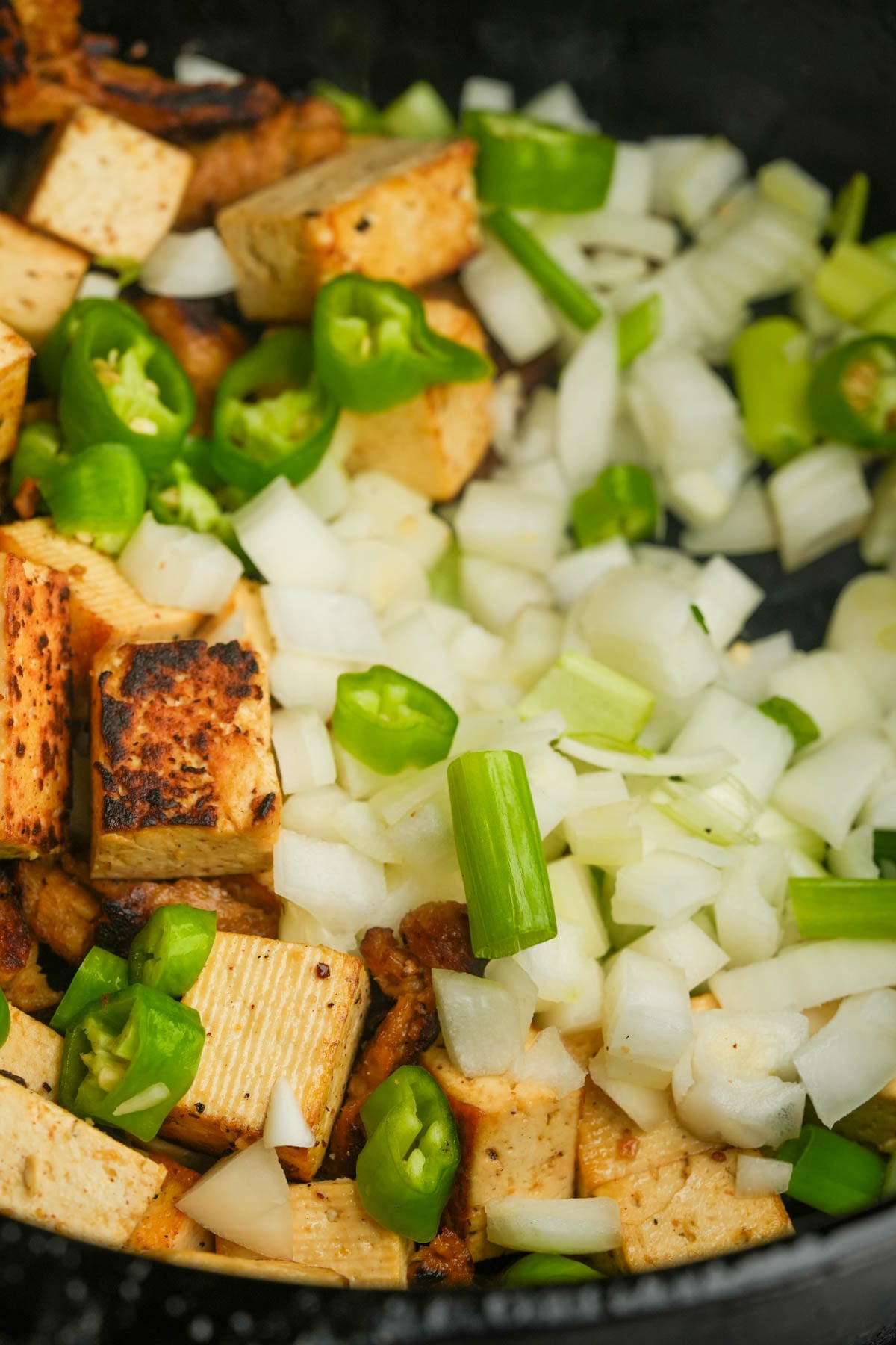 Cubed tofu, chopped onions, and sliced green peppers are being cooked together in a pan.