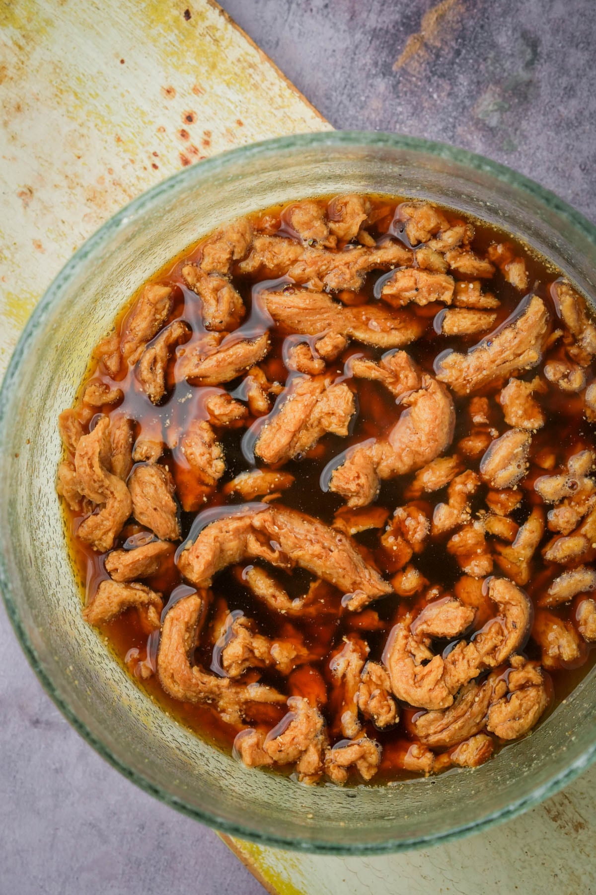 Bits of soy curls are marinating in a reddish-brown sauce inside a glass bowl on a yellow and gray surface.