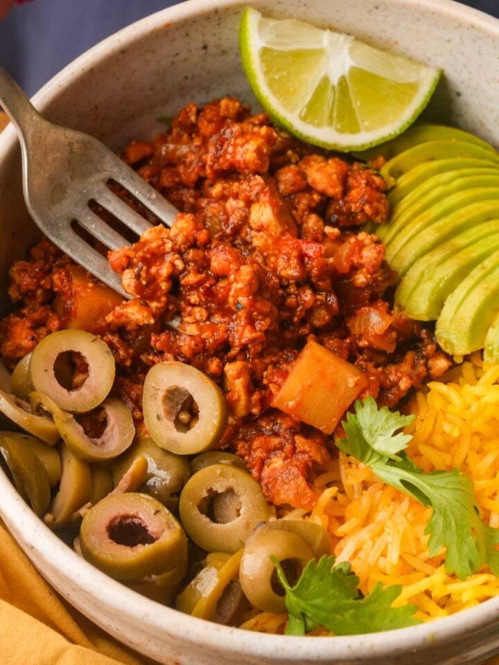 A bowl containing vegan picadillo, sliced avocado, yellow rice, sliced green olives, fresh cilantro, and a lime wedge, with a fork resting inside.