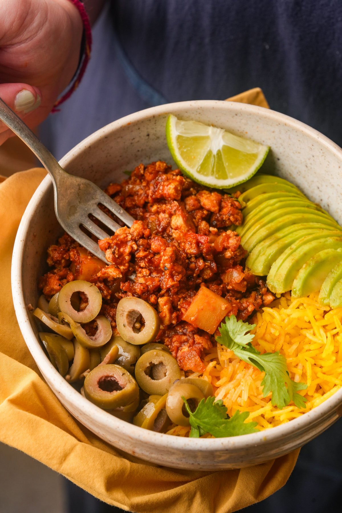 A bowl with yellow rice, sliced avocado, green olives, vegan picadillo, fresh cilantro, and lime wedges, held by a person using a fork.