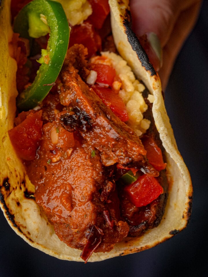 A hand holds a taco filled with saucy vegan carne asada, diced tomatoes, scrambled eggs, and sliced jalapeños in a slightly charred tortilla.