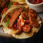A close-up of a vegan carne asada taco with grilled slices of marinated meat, salsa, chopped onions, and cilantro on a lightly charred corn tortilla. Tortilla chips and salsa are in the background.