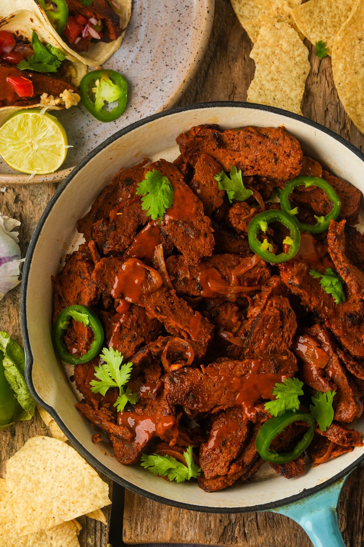 A pan of sliced, saucy vegan carne asada garnished with cilantro and jalapeño, surrounded by tortilla chips, lime wedges, and a plate with loaded tacos.