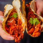 Close-up of two hands holding tacos filled with seasoned vegan carnitas, onions, and cilantro in slightly charred tortillas.