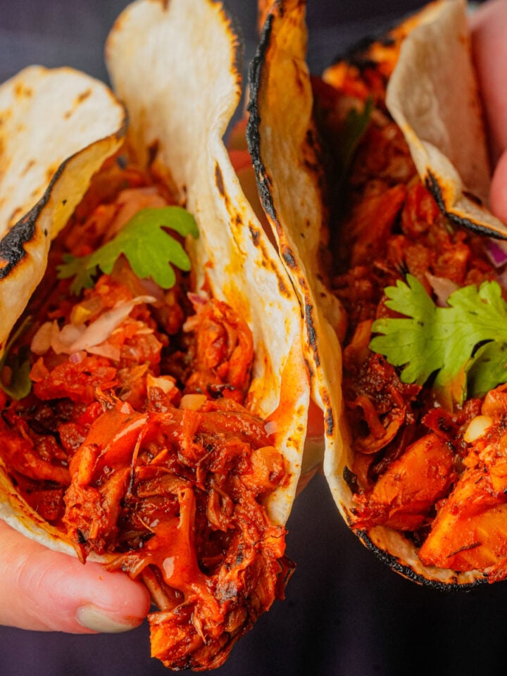 Close-up of two hands holding tacos filled with seasoned vegan carnitas, onions, and cilantro in slightly charred tortillas.