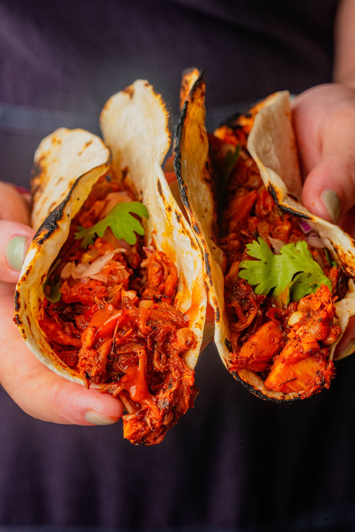 Two hands hold tacos filled with shredded, seasoned meat, garnished with cilantro and onions on lightly charred tortillas.
