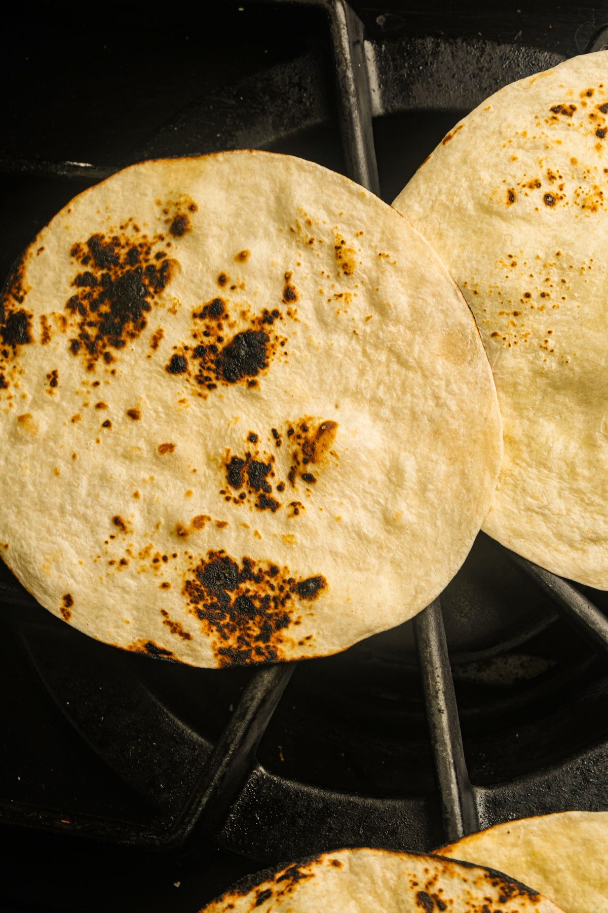 Three corn tortillas with browned, charred spots are warming directly on a gas stove burner.