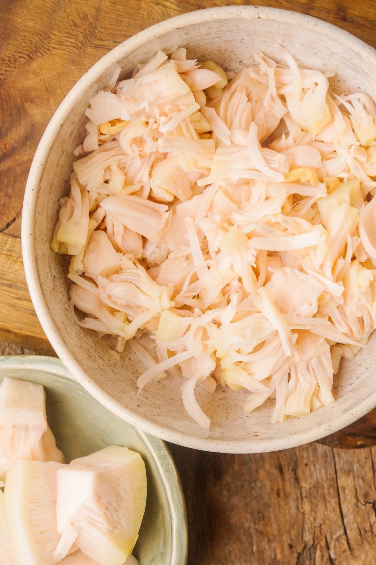 A bowl filled with shredded jackfruit pieces next to another bowl containing larger chunks of jackfruit on a wooden surface.