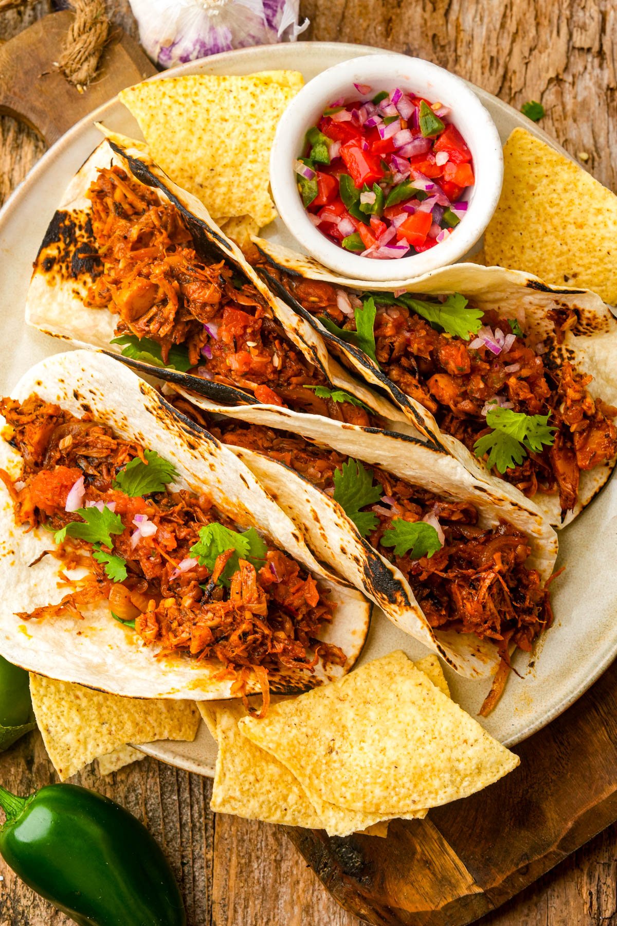 Three grilled vegan carnitas tacos filled with shredded jackfruit and cilantro, served with tortilla chips and a small bowl of salsa on a rustic wooden surface.