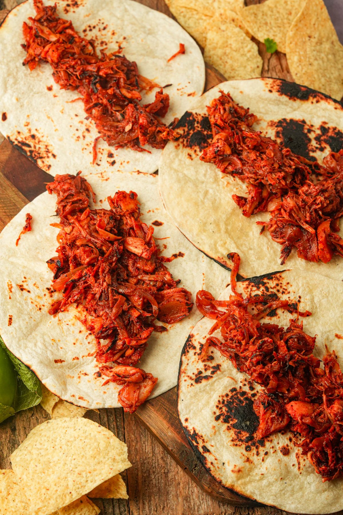 Four flour tortillas with shredded vegan carnitas are arranged on a wooden board, surrounded by tortilla chips.
